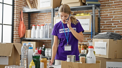 Young blonde woman volunteer writing on clipboard talking on smartphone at charity center