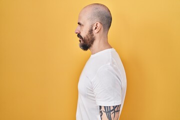 Young hispanic man with tattoos standing over yellow background looking to side, relax profile pose...