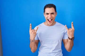 Young hispanic man standing over blue background shouting with crazy expression doing rock symbol with hands up. music star. heavy music concept.