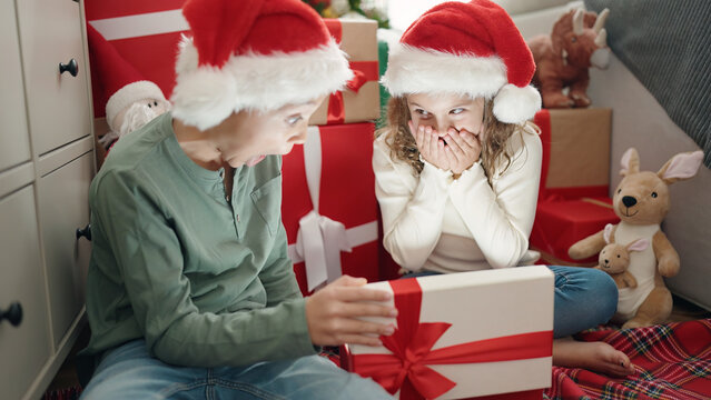 Two kids unpacking gift sitting on floor by christmas tree at home