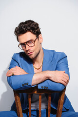 Confident and stylish model in eyeglasses, blue suit and golden chain sitting on chair on grey