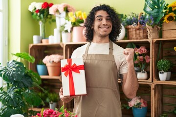 Hispanic man with curly hair working at florist shop holding gift screaming proud, celebrating victory and success very excited with raised arm