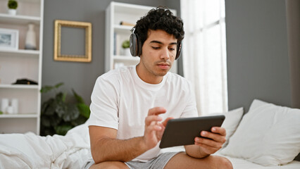 Young latin man using touchpad sitting on the bed wearing headphones at bedroom