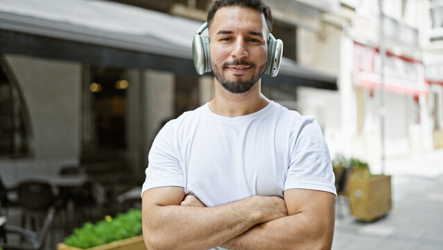 Young arab man listening to music standing with arms crossed gesture at coffee shop terrace