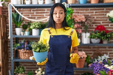 Hispanic woman working at florist shop holding plant puffing cheeks with funny face. mouth inflated with air, catching air. - obrazy, fototapety, plakaty