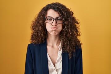 Hispanic woman with curly hair standing over yellow background puffing cheeks with funny face. mouth inflated with air, crazy expression. - obrazy, fototapety, plakaty