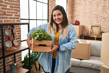 Young hispanic woman moving to a new home holding plants looking positive and happy standing and smiling with a confident smile showing teeth