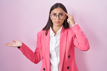 Young hispanic woman wearing business clothes and glasses confused and annoyed with open palm showing copy space and pointing finger to forehead. think about it.