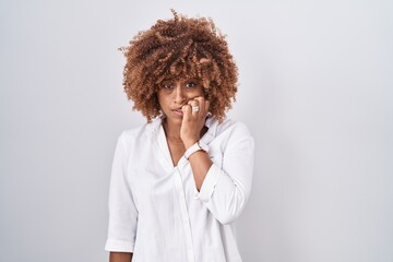 Young hispanic woman with curly hair standing over white background looking stressed and nervous with hands on mouth biting nails. anxiety problem.