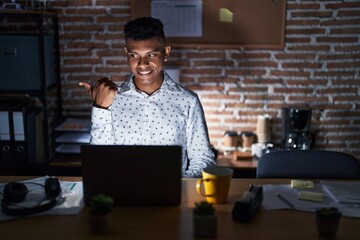 Young hispanic man working at the office at night smiling with happy face looking and pointing to...