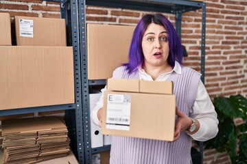 Plus size woman wit purple hair working at small business ecommerce holding boxes in shock face,...