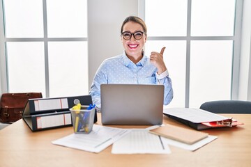 Young hispanic woman working at the office wearing glasses smiling happy and positive, thumb up doing excellent and approval sign