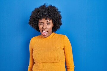 Fototapeta na wymiar Black woman with curly hair standing over blue background winking looking at the camera with sexy expression, cheerful and happy face.