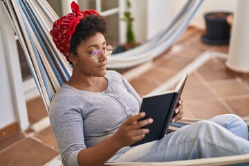 African american woman reading book lying on hammock at home terrace