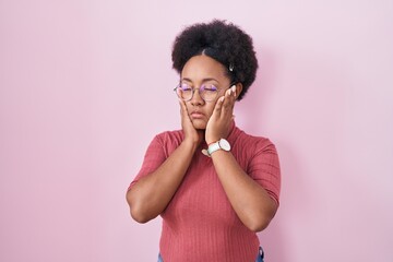 Beautiful african woman with curly hair standing over pink background tired hands covering face,...