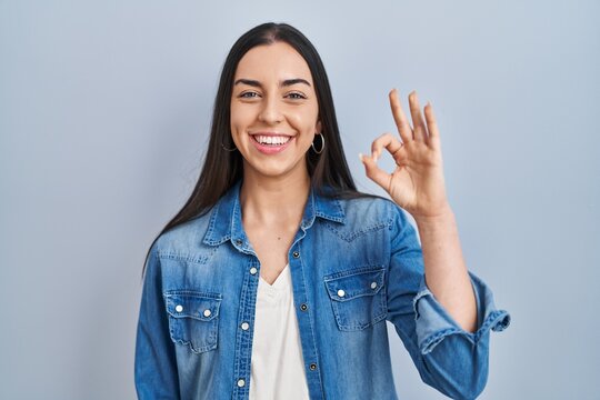 Hispanic woman standing over blue background smiling positive doing ok sign with hand and fingers. successful expression.