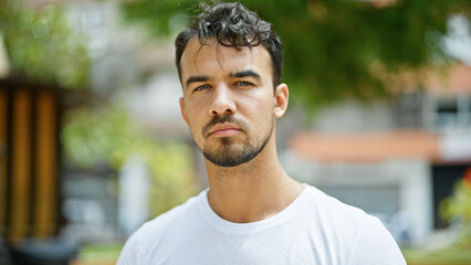 Young hispanic man standing with serious expression at park - obrazy, fototapety, plakaty