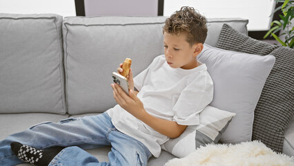 Adorable blond boy, seriously engrossed in watching a video on his smartphone, eating a sweet...