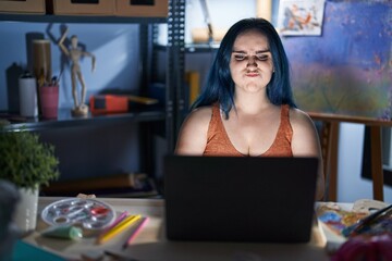 Young modern girl with blue hair sitting at art studio with laptop at night puffing cheeks with funny face. mouth inflated with air, crazy expression. - obrazy, fototapety, plakaty