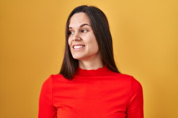 Young hispanic woman standing over yellow background looking away to side with smile on face, natural expression. laughing confident.