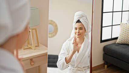 Young beautiful hispanic woman wearing bathrobe touching face looking on mirror at bedroom