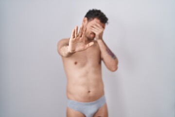 Fototapeta premium Young hispanic man standing shirtless wearing underware covering eyes with hands and doing stop gesture with sad and fear expression. embarrassed and negative concept.