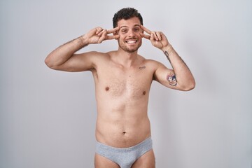 Fototapeta premium Young hispanic man standing shirtless wearing underware doing peace symbol with fingers over face, smiling cheerful showing victory