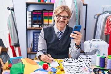 Young blond man tailor using smartphone writing on notebook at clothing factory