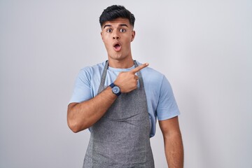 Hispanic young man wearing apron over white background surprised pointing with finger to the side, open mouth amazed expression.