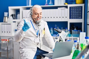 Middle age man with grey hair working at scientist laboratory doing video call pointing thumb up to the side smiling happy with open mouth