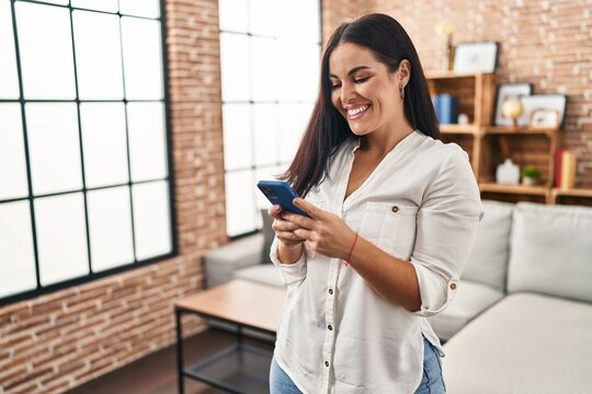 Young beautiful hispanic woman using smartphone standing at home