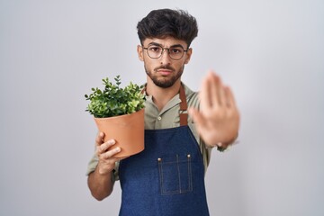 Arab man with beard holding green plant pot doing stop sing with palm of the hand. warning...