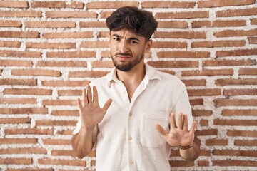 Arab man with beard standing over bricks wall background moving away hands palms showing refusal and denial with afraid and disgusting expression. stop and forbidden.