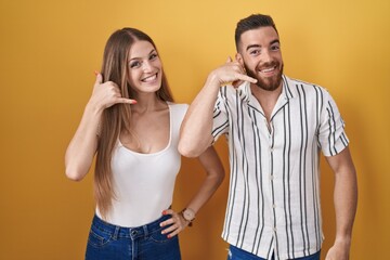 Young couple standing over yellow background smiling doing phone gesture with hand and fingers like talking on the telephone. communicating concepts.