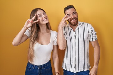 Young couple standing over yellow background doing peace symbol with fingers over face, smiling cheerful showing victory