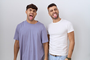 Homosexual gay couple standing over white background sticking tongue out happy with funny...