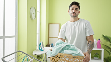 Young hispanic man holding basket with clothes with sad expression at laundry room