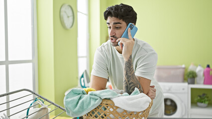 Young hispanic man talking on smartphone leaning on basket with clothes at laundry room