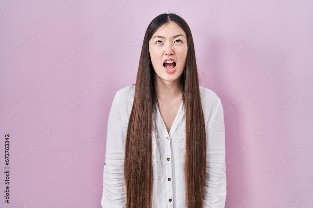 Sticker Chinese young woman standing over pink background angry and mad screaming frustrated and furious, shouting with anger. rage and aggressive concept.