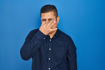 Hispanic young man standing over blue background smelling something stinky and disgusting, intolerable smell, holding breath with fingers on nose. bad smell