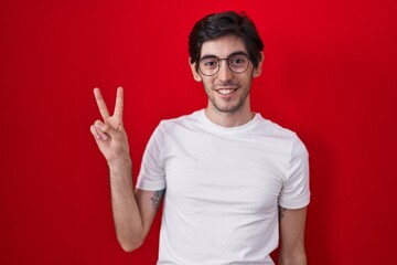 Young hispanic man standing over red background smiling with happy face winking at the camera doing...