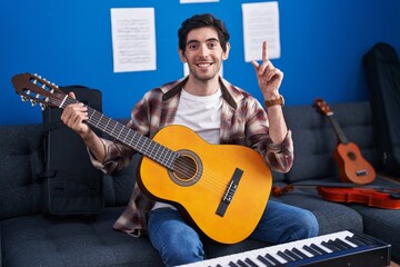 Young hispanic man playing classic guitar at music studio surprised with an idea or question pointing finger with happy face, number one