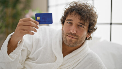 Young hispanic man wearing bathrobe holding credit card at bedroom