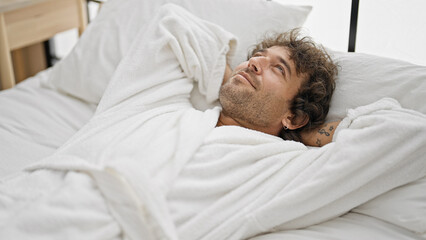 Young hispanic man wearing bathrobe relaxed on bed at bedroom