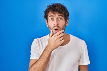 Hispanic young man standing over blue background looking fascinated with disbelief, surprise and amazed expression with hands on chin