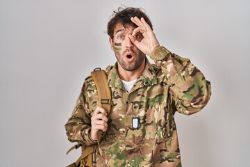 Hispanic young man wearing camouflage army uniform doing ok gesture shocked with surprised face, eye looking through fingers. unbelieving expression.