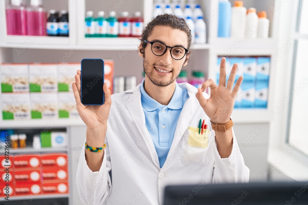 Poster Young hispanic man working at pharmacy drugstore showing smartphone screen doing ok sign with fingers, smiling friendly gesturing excellent symbol