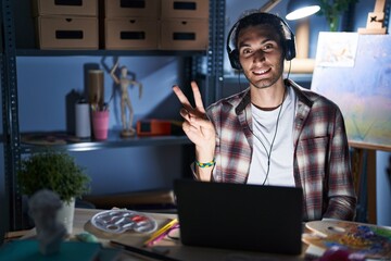 Young hispanic man sitting at art studio with laptop late at night smiling with happy face winking at the camera doing victory sign. number two. - obrazy, fototapety, plakaty