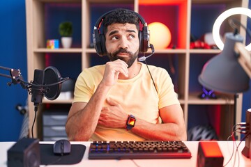 Hispanic man with beard playing video games with headphones with hand on chin thinking about...