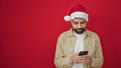 Young hispanic man wearing christmas hat using smartphone over isolated red background
